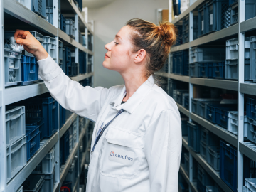 Laboratorijska tehničarka u Eurofins skladištu za stabilnost, okružen policama ispunjenim materijalima. Tehničar pažljivo pregleda sadržaj jednog od spremnika, naglašavajući preciznost i pažnju na detalje u postupcima testiranja stabilnosti.