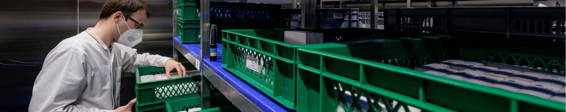 Tehničar koji nosi zaštitnu masku i zaštitne naočale unutar prostorije za skladištenje i stabilnost u Eurofins BioPharma Product Testing, stavlja zelenu kutiju s materijalima na policu.