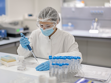 Istraživačica u laboratoriju, odjevena u bijeli mantil, rukavice, zaštitnu masku i zaštitnu mrežicu za kosu, koristi pipetu za prijenos tekućine u posudu, sprovodeći testiranje molekularne biologije. Radno mjesto ima epruvete, laboratorijske alate i druge znanstvene materijale.