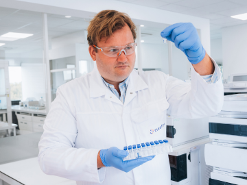 Laboratorijski tehničar u laboratorijskom mantilu, zaštitnim naočalama i plavim rukavicama, drži bočicu dok provodi fizikalno-kemijske i mikrobiološke analize. Scena naglašava pažljivost tehničara u izvođenju znanstvenih testova u kontroliranom laboratorijskom okruženju Eurofinsa.